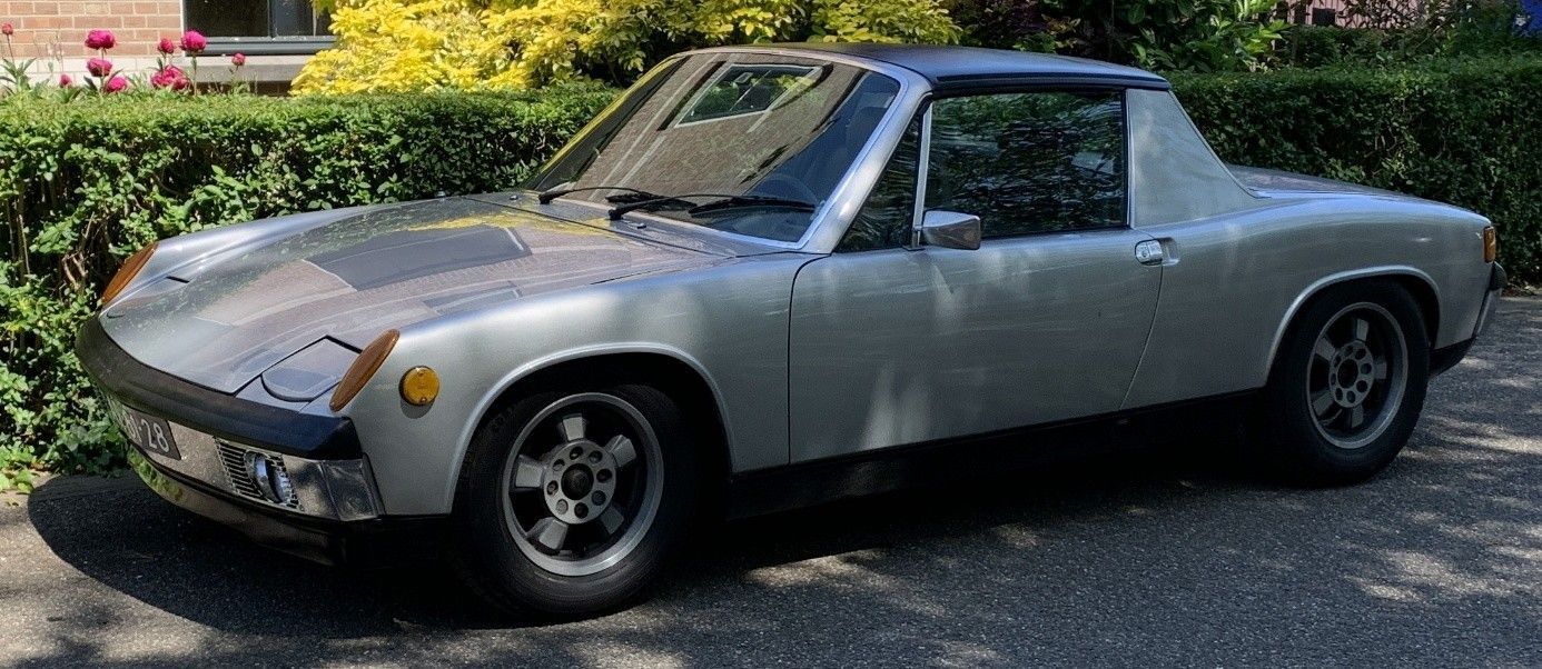 Een zilverkleurige Porsche 914 staat geparkeerd op een geplaveide oprit met groen gebladerte op de achtergrond.