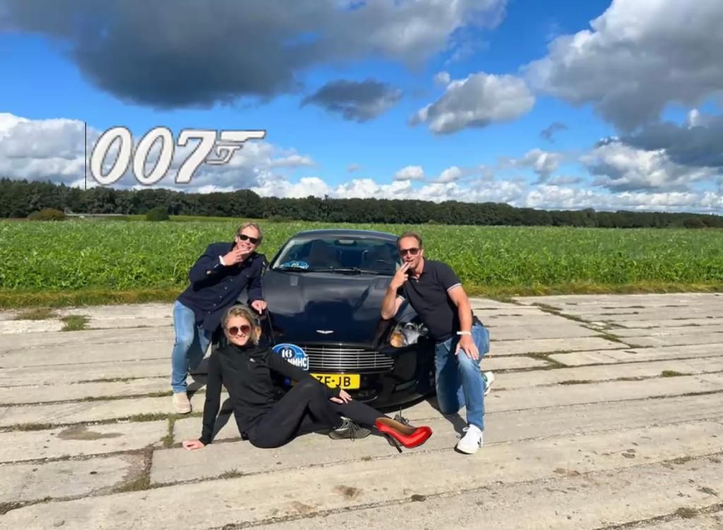 Drie mensen poseren met een zwarte auto op een betonnen pad; het 