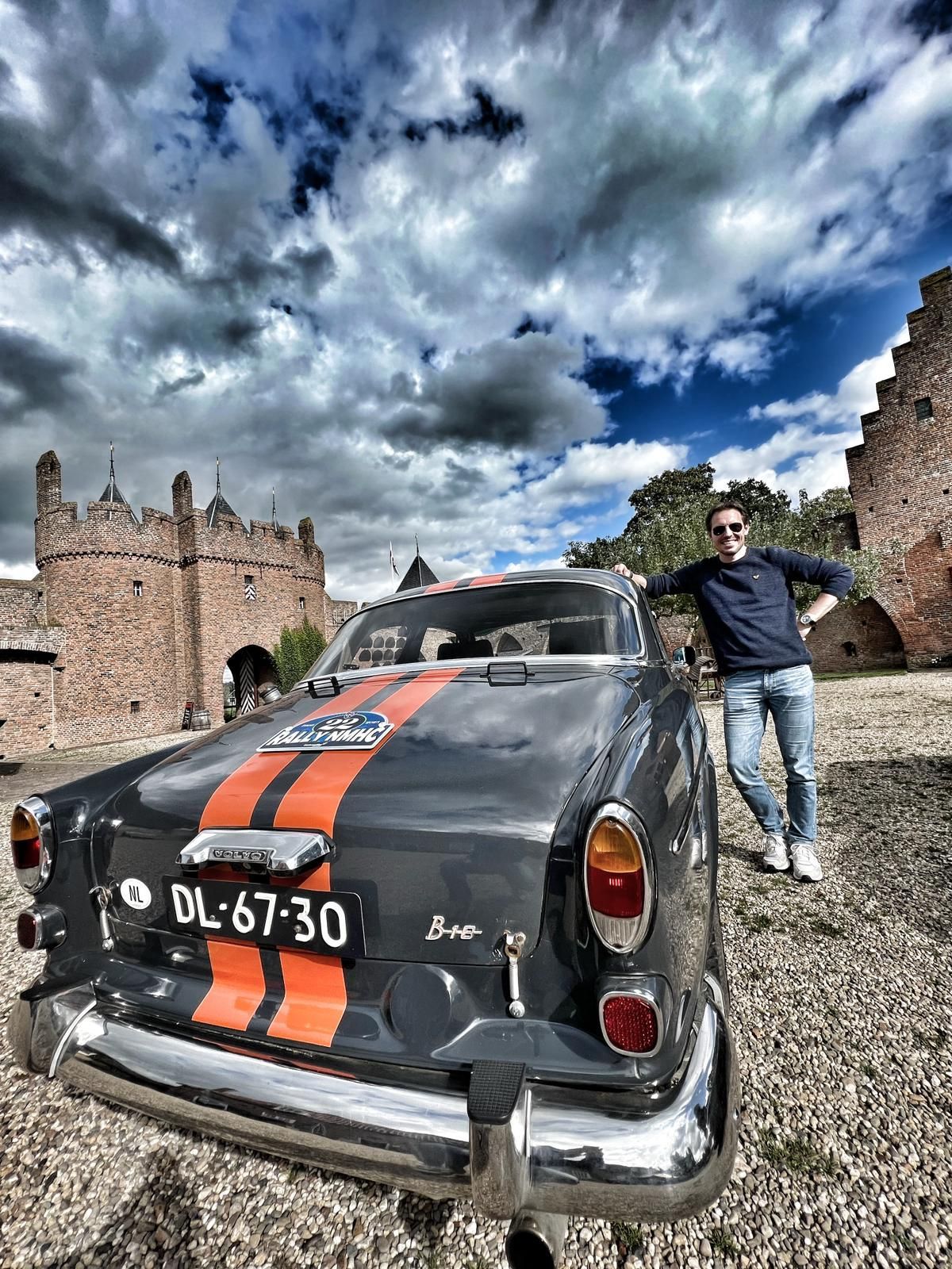 Een man poseert naast een vintage grijze Volvo met oranje strepen voor een kasteel onder een bewolkte hemel.