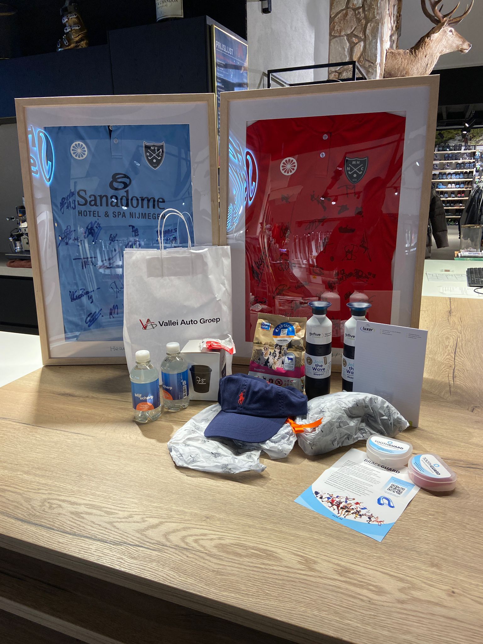 Presentatie van handelswaar: ingelijste shirts, drankjes en accessoires op een houten tafel, in een winkelomgeving.