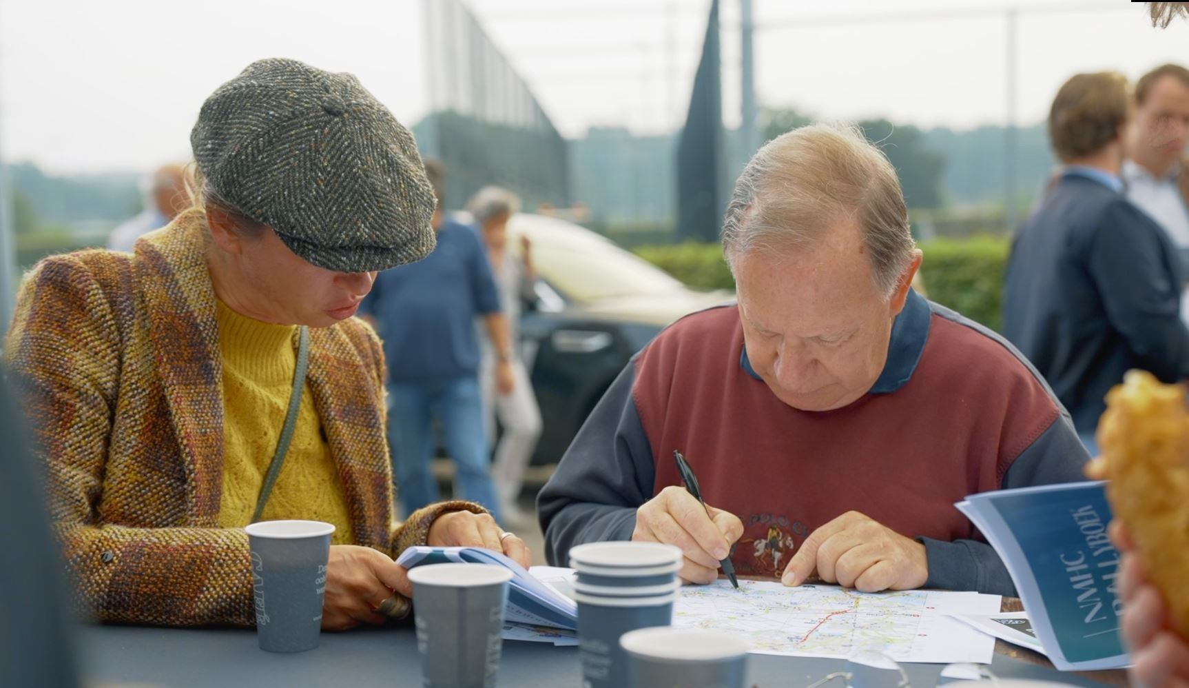 Twee mannen zitten aan een tafel en bekijken papieren kaarten, met anderen op de achtergrond. Kopjes en een snack zijn zichtbaar.