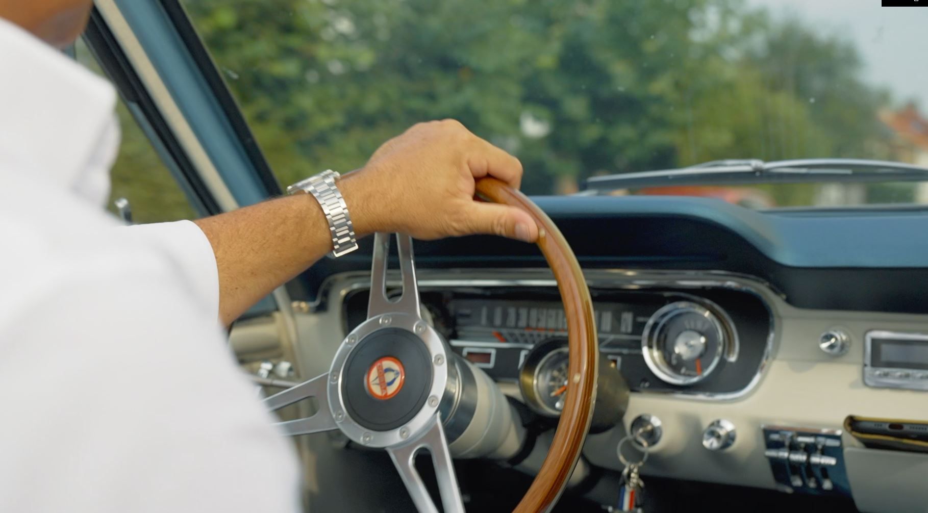 De hand van de bestuurder op het stuur van een klassieke blauwe auto, het dashboard is zichtbaar.