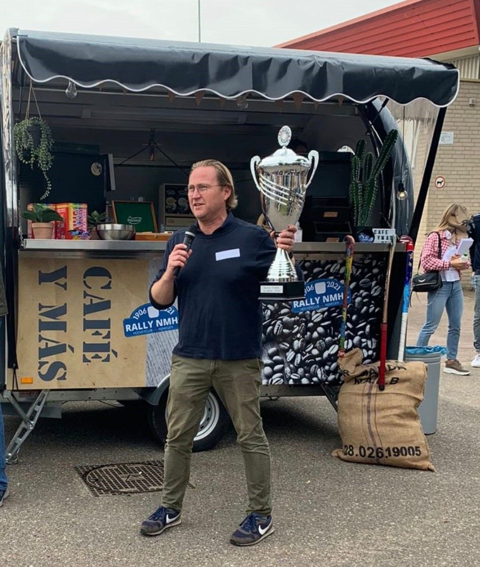 Een man met een trofee in zijn handen spreekt in een microfoon voor een eetkraam, in een buitenomgeving.