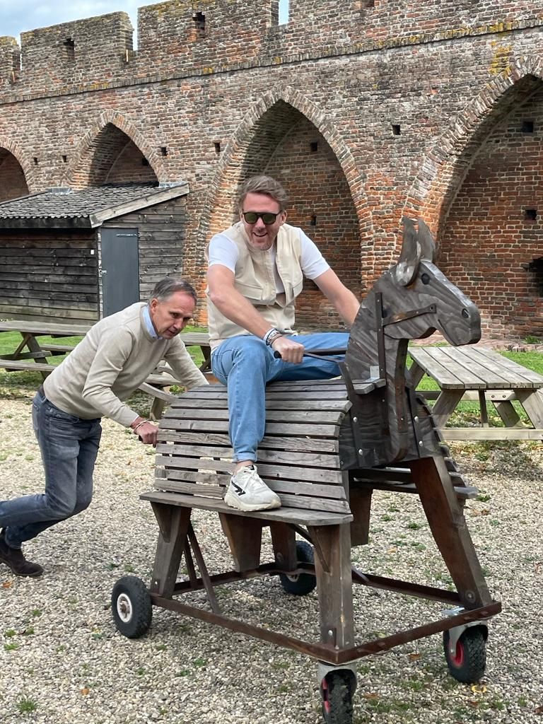 Twee mannen poseren met een houten paard op wielen bij een bakstenen gebouw. ​​De ene zit op het paard, de andere duwt het voort.