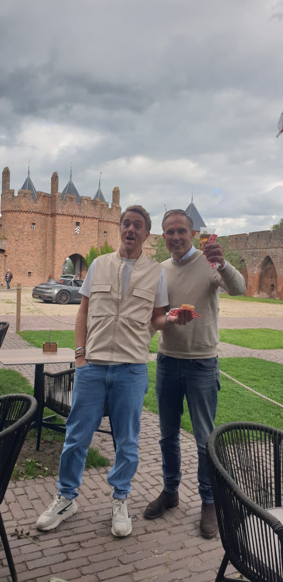 Twee mannen poseren voor een foto buiten voor een bakstenen kasteel. Een van hen houdt een drankje omhoog. Bewolkte lucht.