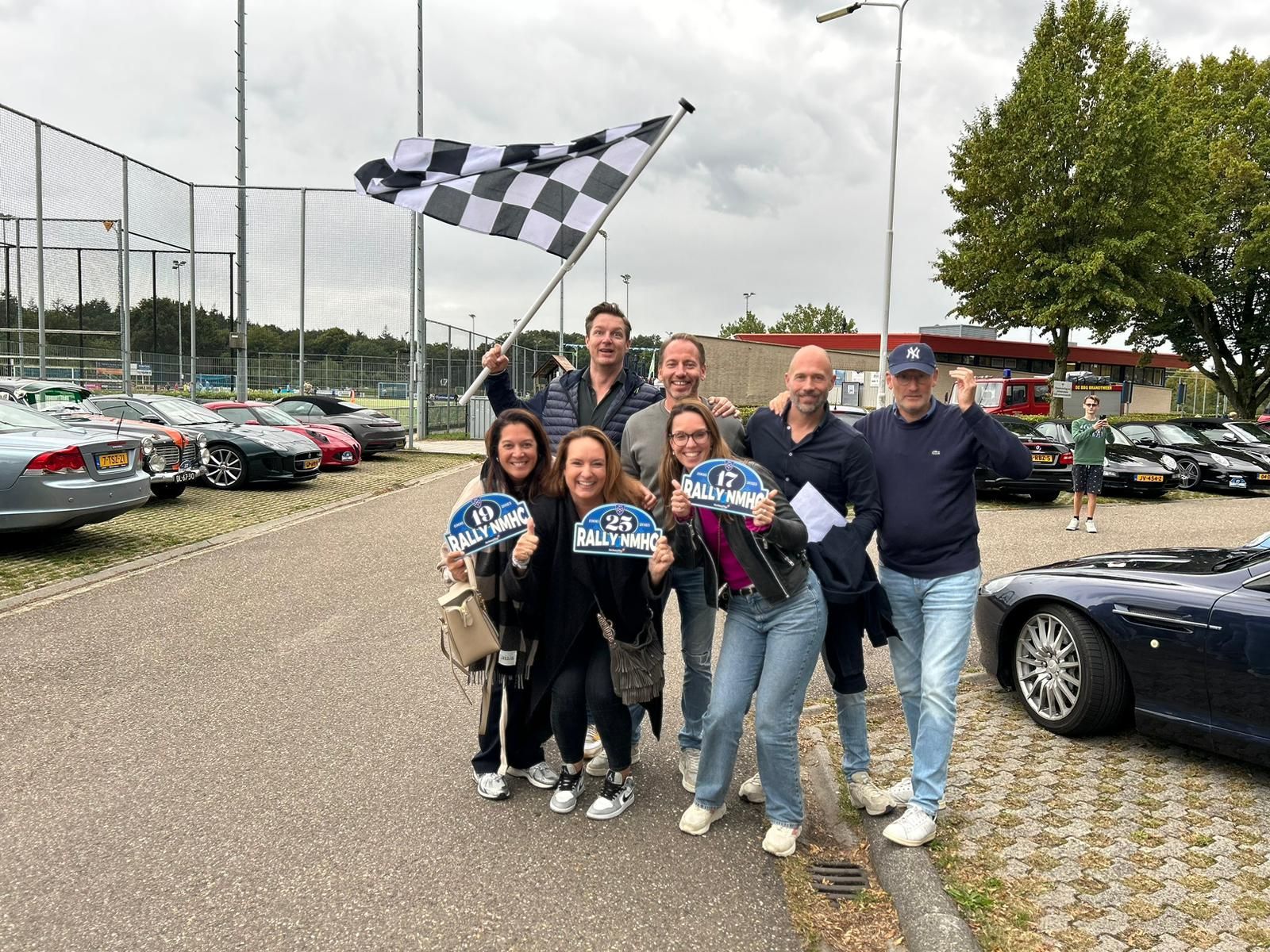 Een groep mensen viert feest naast geparkeerde auto's; iemand zwaait met een geblokte vlag.