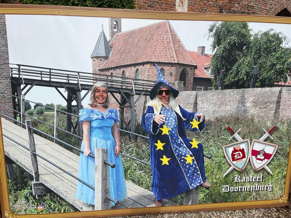 Twee mensen in kostuums poseren op een brug voor een kasteel. De ene draagt ​​een blauwe jurk, de andere een tovenaarskostuum.