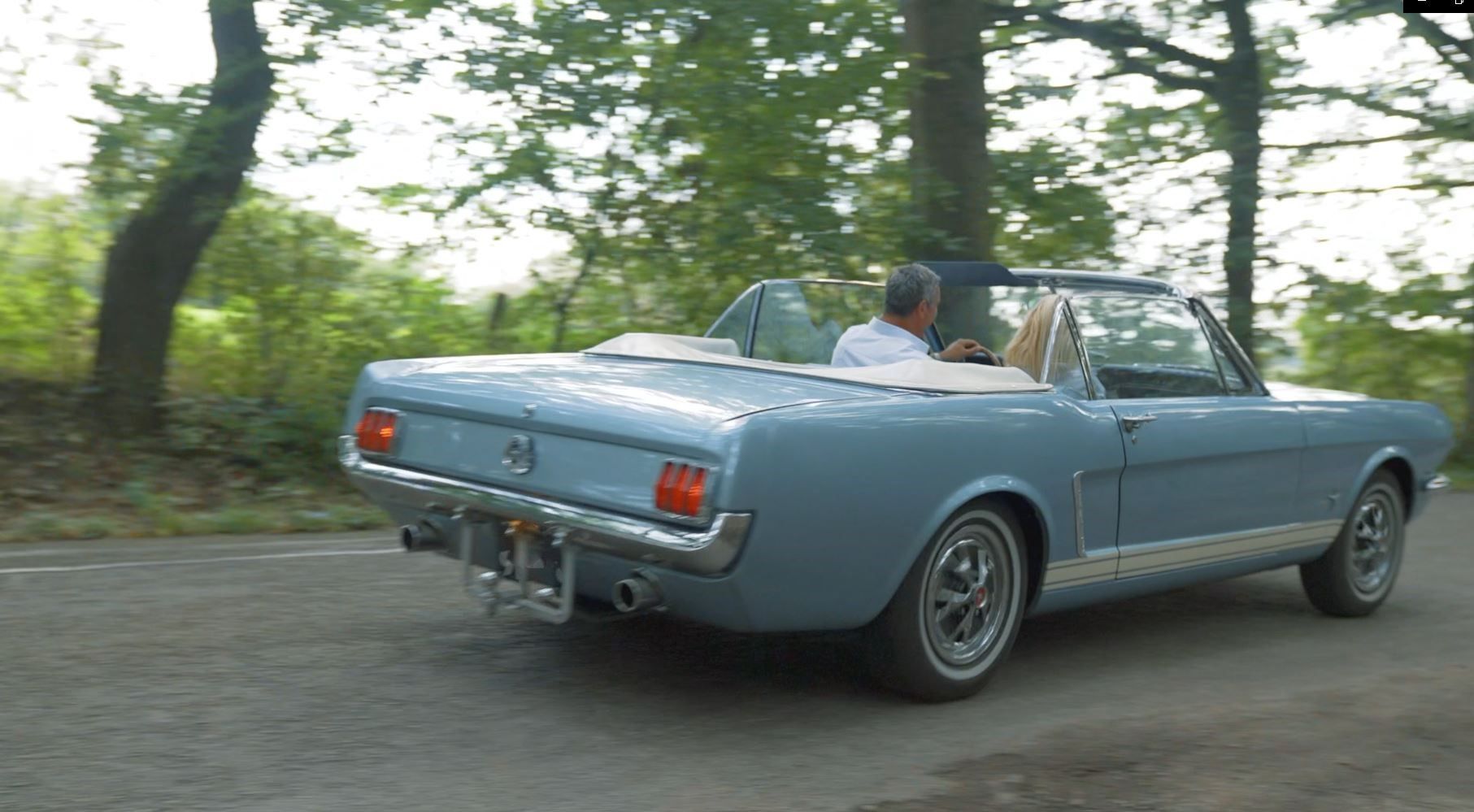 Een lichtblauwe klassieke cabriolet rijdt over een met bomen omzoomde weg.
