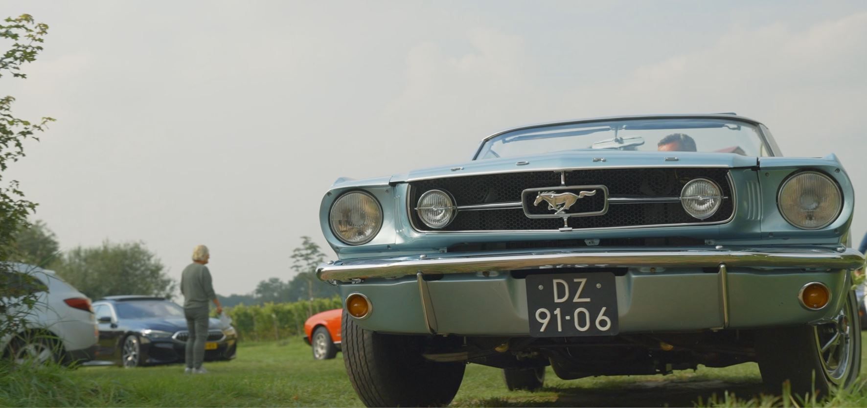 Een blauwe cabriolet Mustang met een Nederlands kenteken staat geparkeerd op het gras. Een persoon staat bij andere auto's.