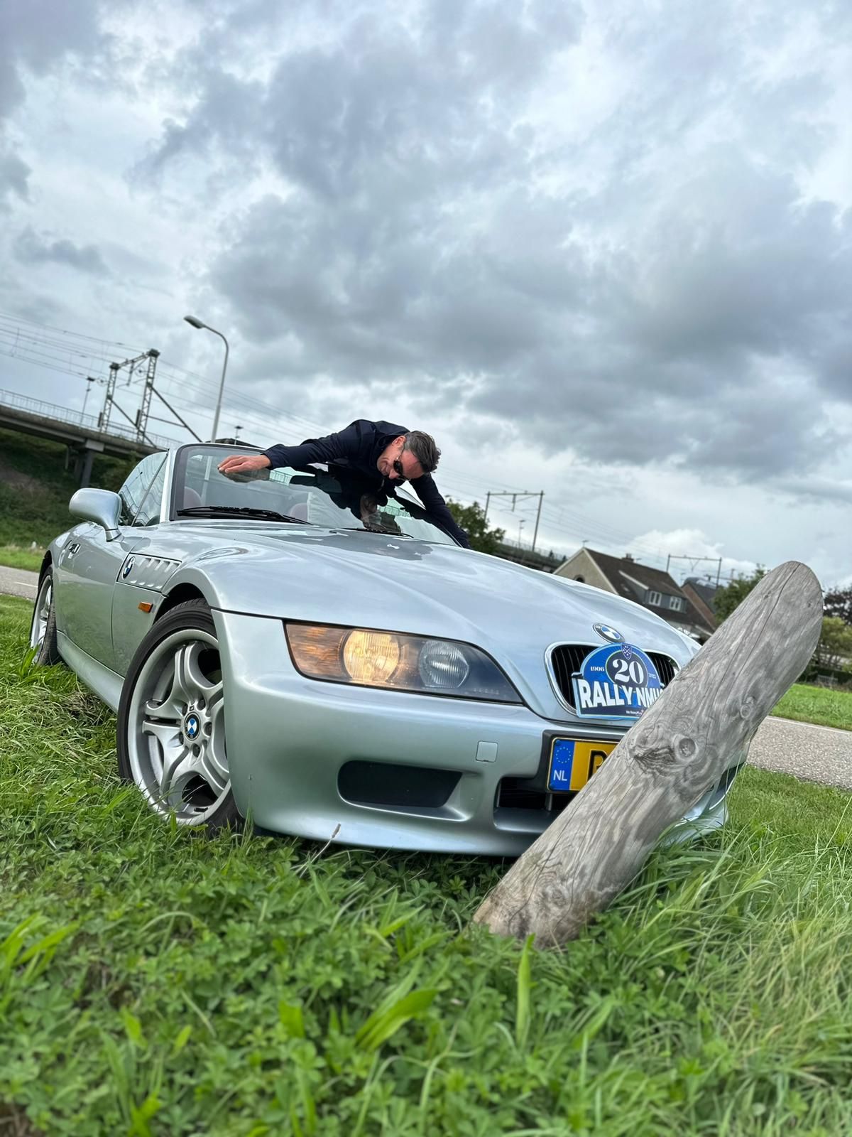 Een man leunt tegen de motorkap van een zilverkleurige cabriolet, die geparkeerd staat in het gras, onder een bewolkte hemel.