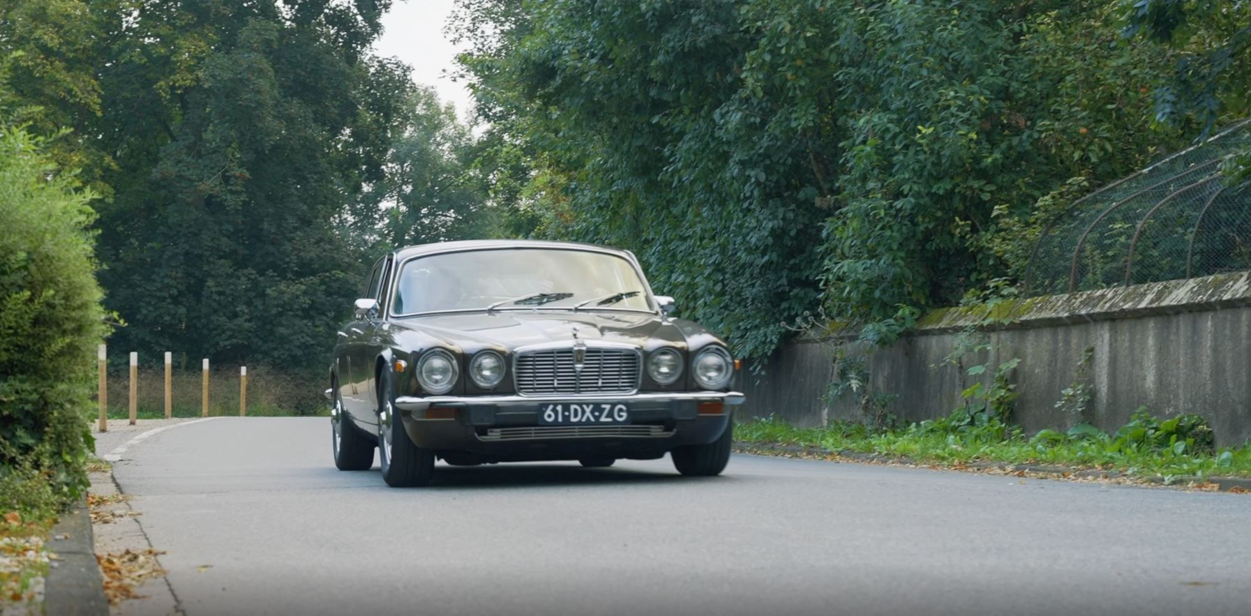 Een klassieke, donkere auto rijdt over een bochtige weg, met bomen en een hek op de achtergrond.