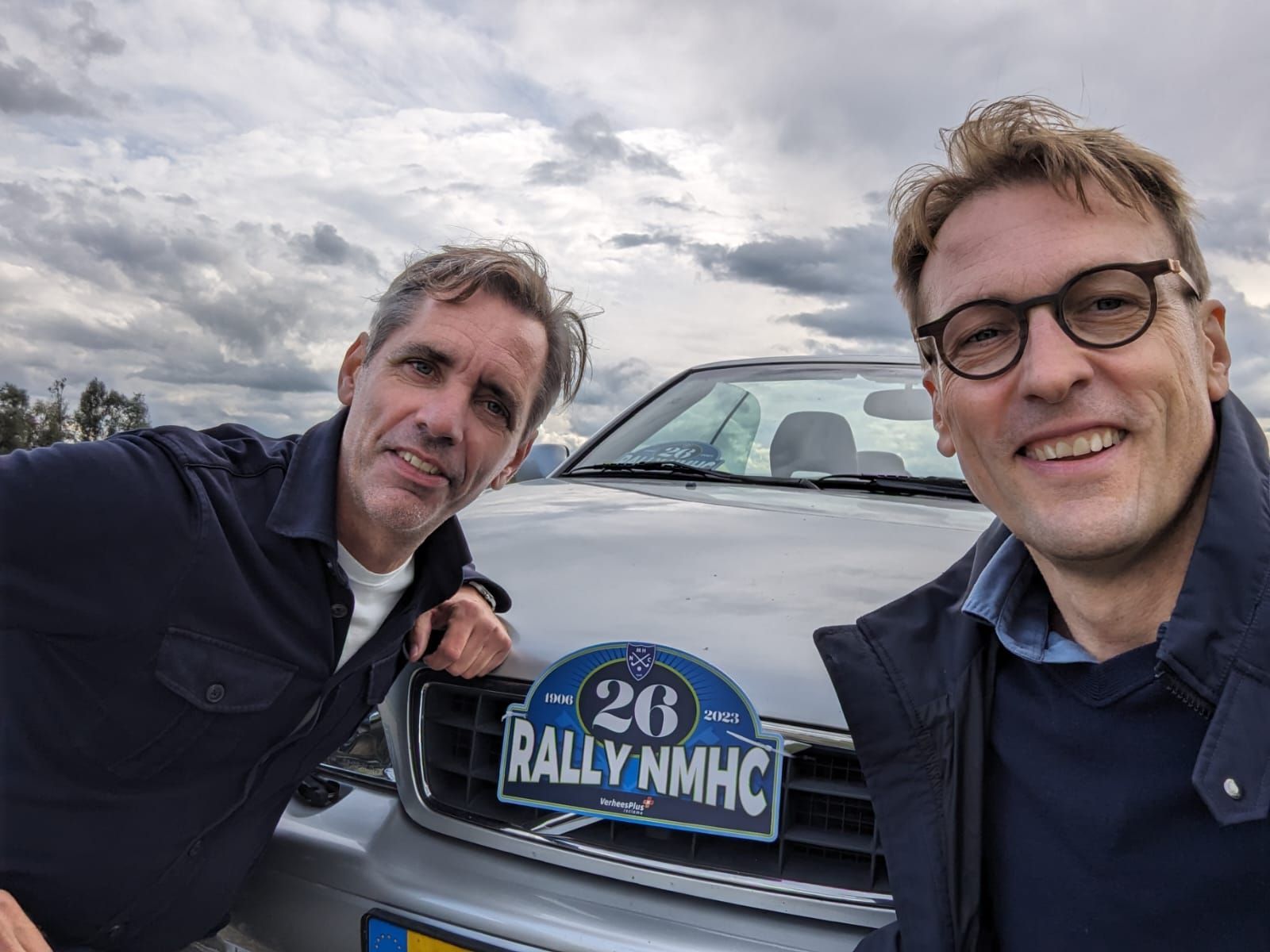 Twee lachende mannen poseren voor een auto met een rallysticker onder een bewolkte hemel.