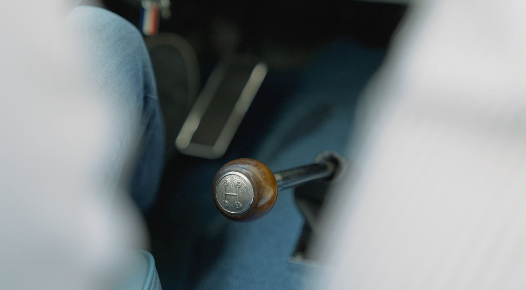 Close-up van de benen van een persoon in een blauwe spijkerbroek, met de focus op een houten versnellingspook en de pedalen van de auto.