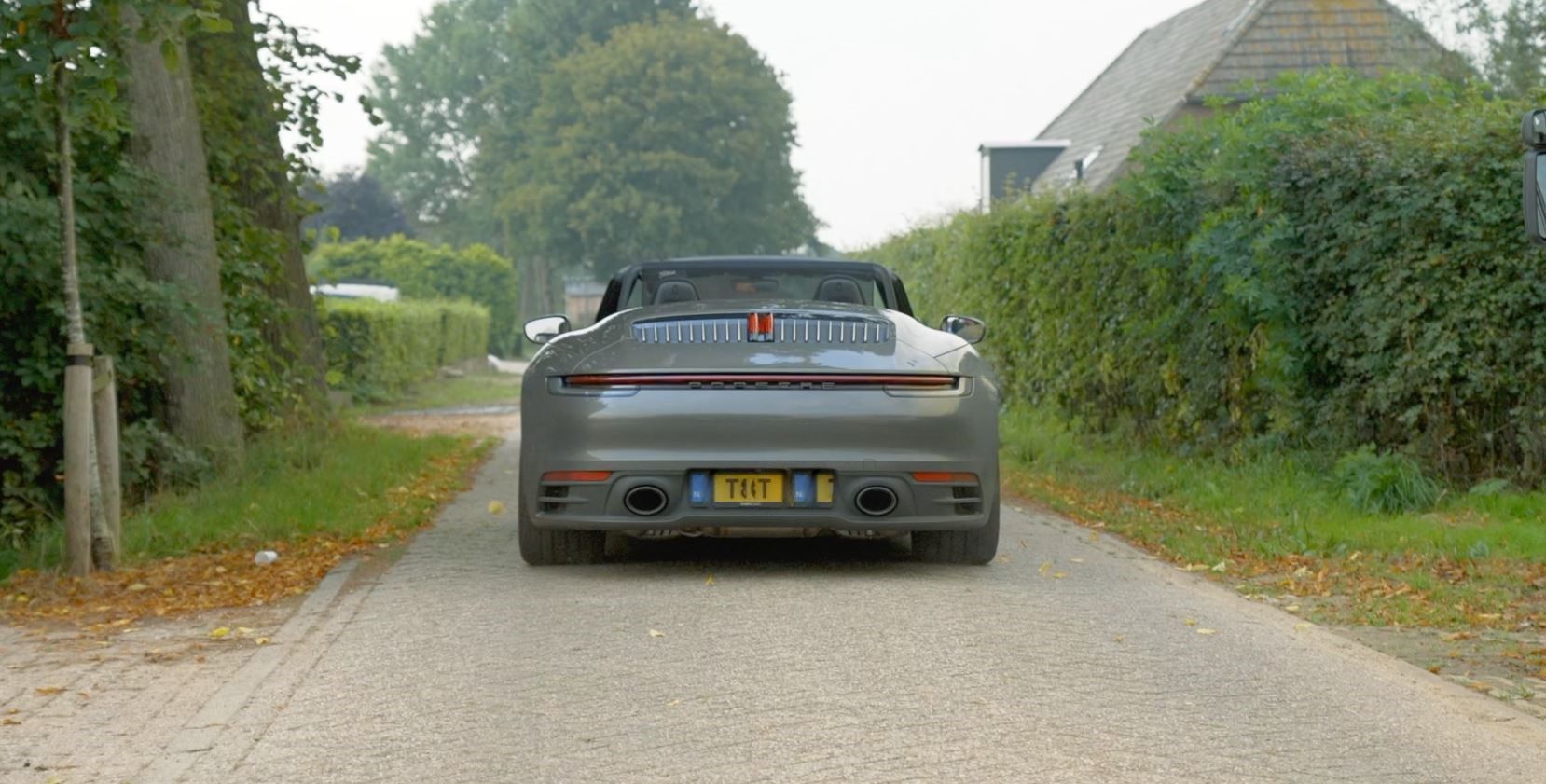 Een grijze Porsche cabriolet rijdt weg over een onverharde weg, langs heggen en bomen.