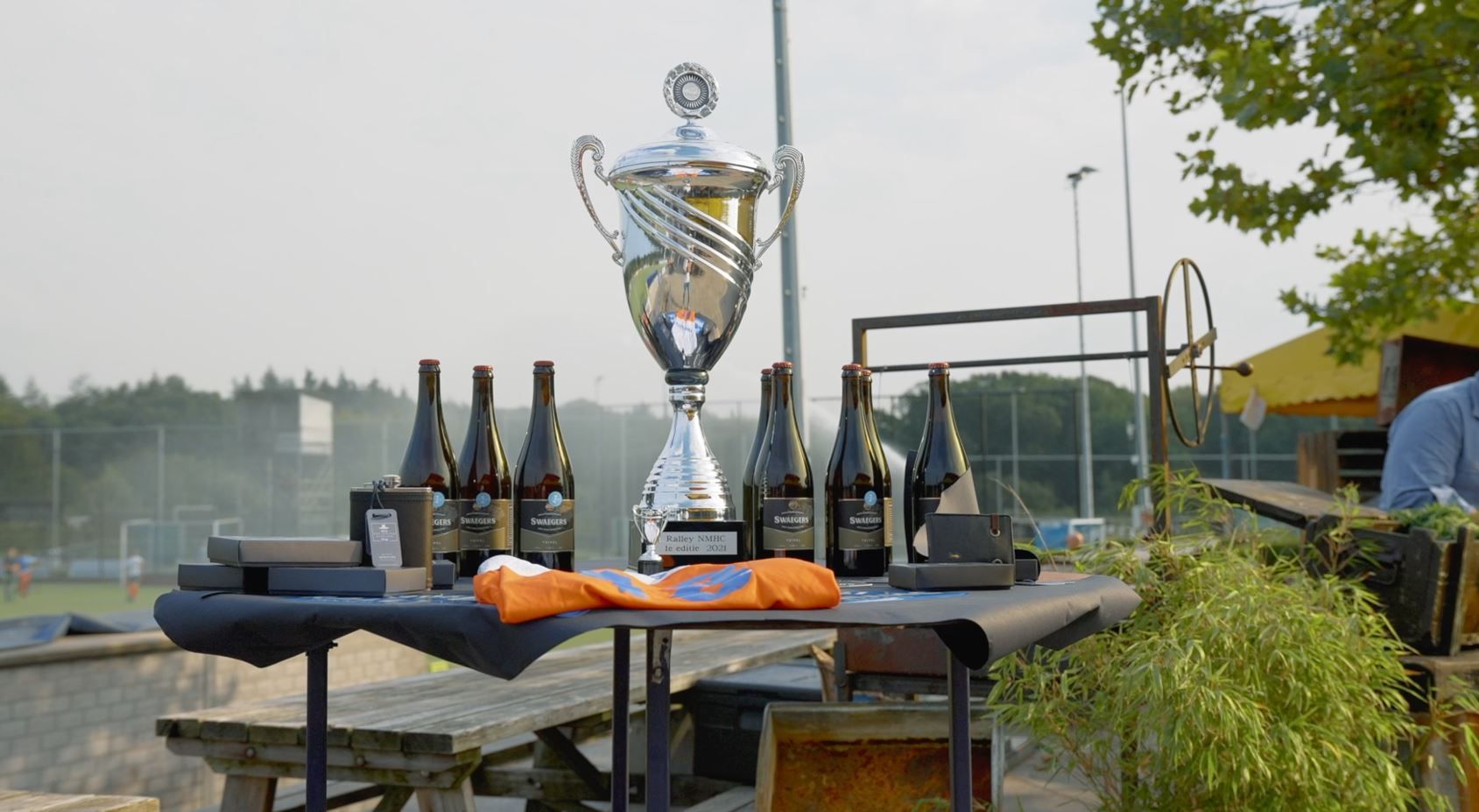 Een zilveren trofee en flessen wijn op een tafel, mogelijk voor een prijsuitreiking, in de buitenlucht.