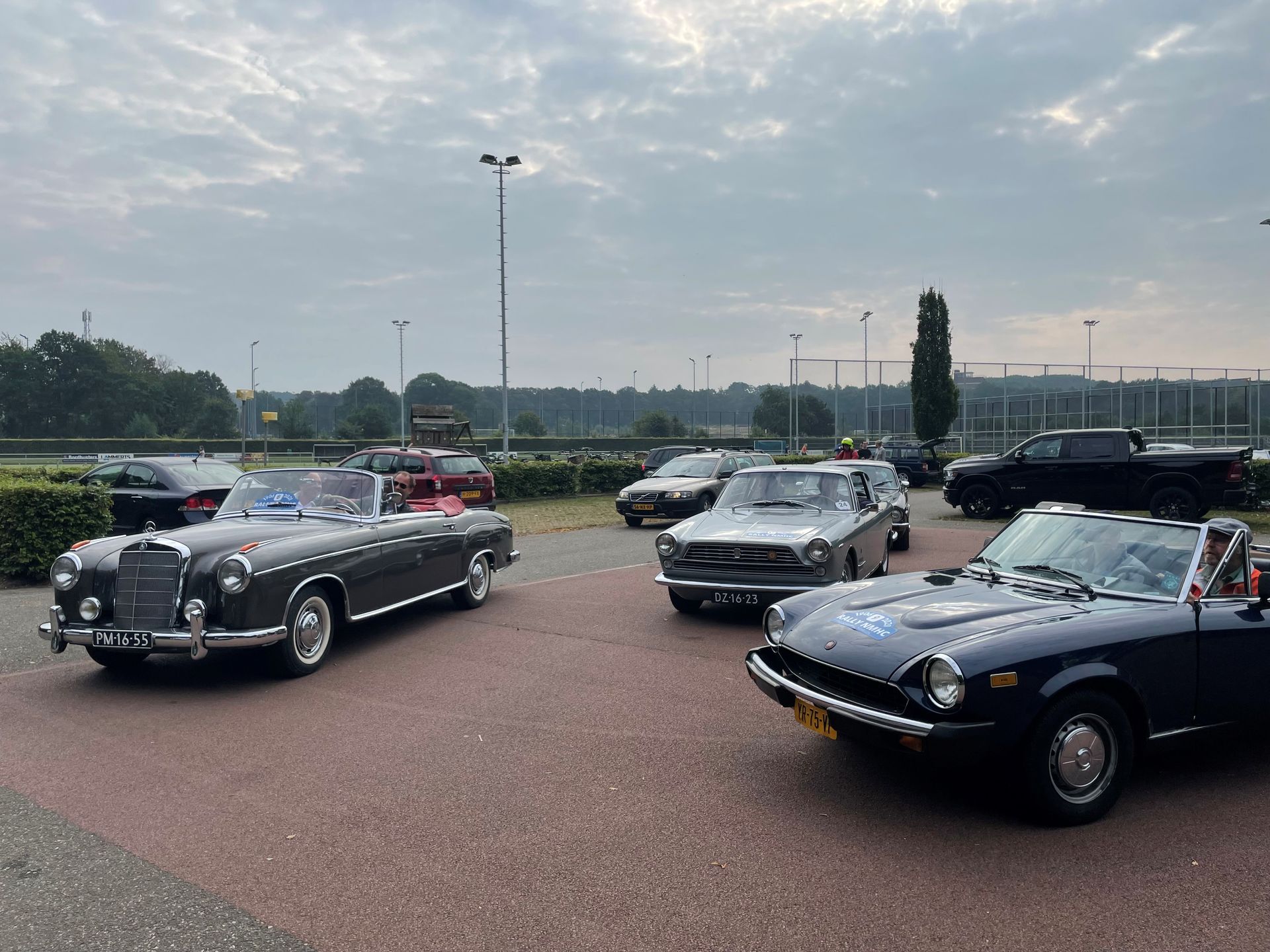 Klassieke auto's staan ​​buiten geparkeerd, waaronder een Mercedes cabriolet en een Triumph. Grijze, bewolkte lucht.