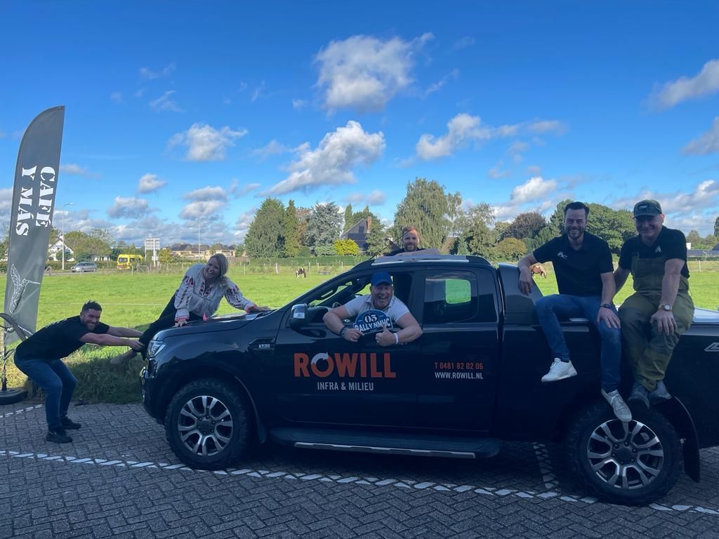 Een groep mensen poseert buiten bij een zwarte pick-up truck. Sommigen leunen ertegenaan en lachen. Op de achtergrond is een bewolkte lucht en een grasveld te zien.