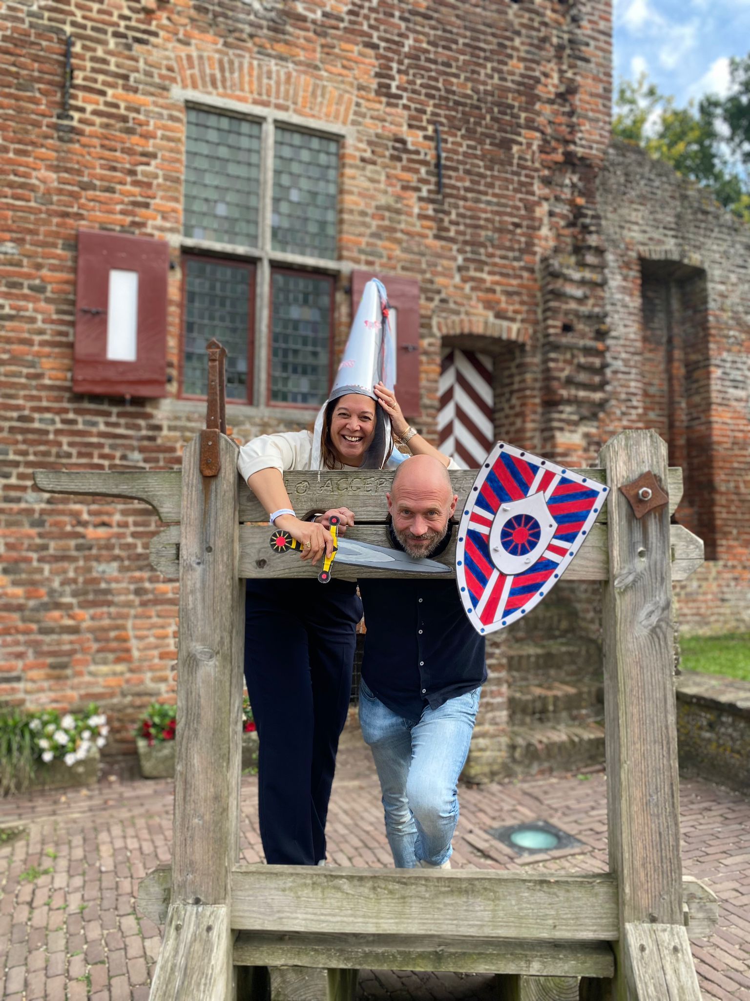 Twee mensen in de schandpaal. Een vrouw met een ezelsmuts houdt een speelgoedzwaard vast, een man een schild, voor een bakstenen kasteel.