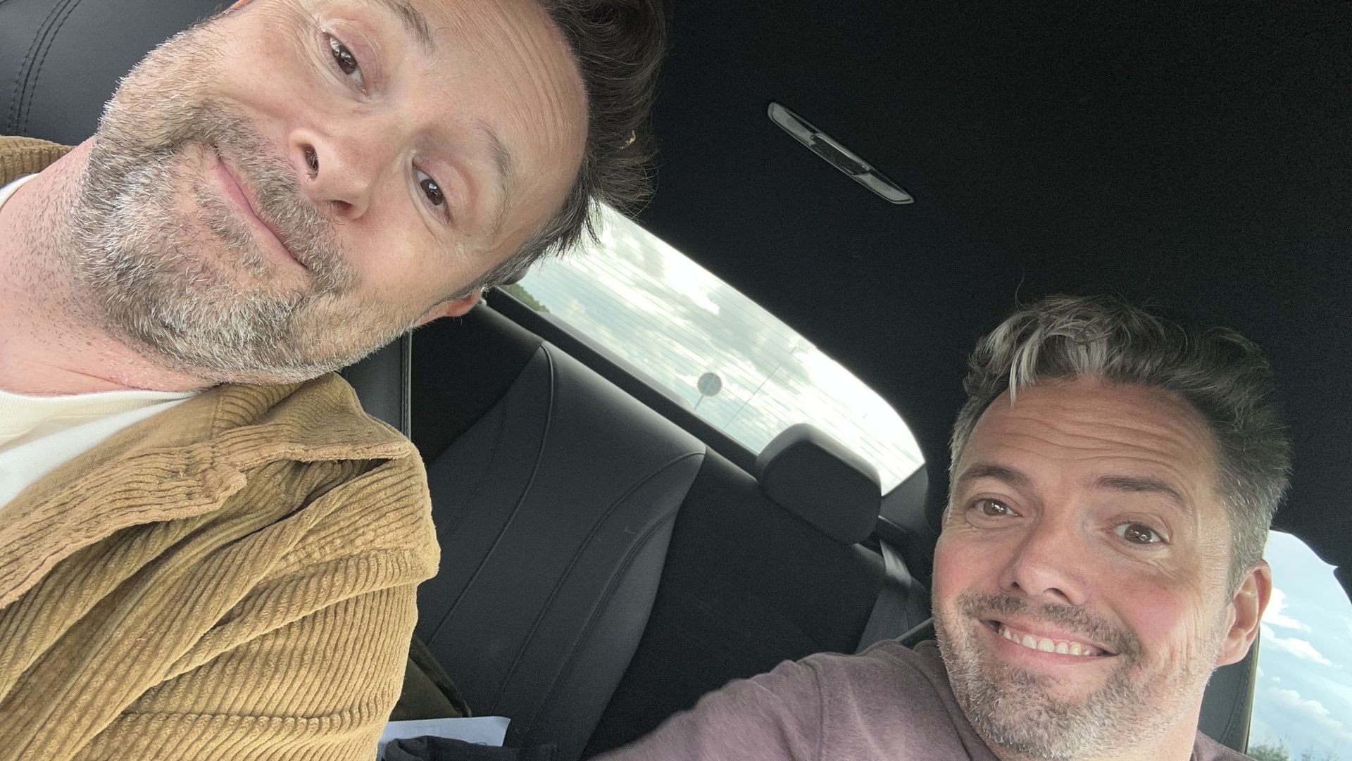 Twee mannen glimlachen in een auto, de een in een bruine jas, de ander in een paars shirt. Buiten schijnt de zon.