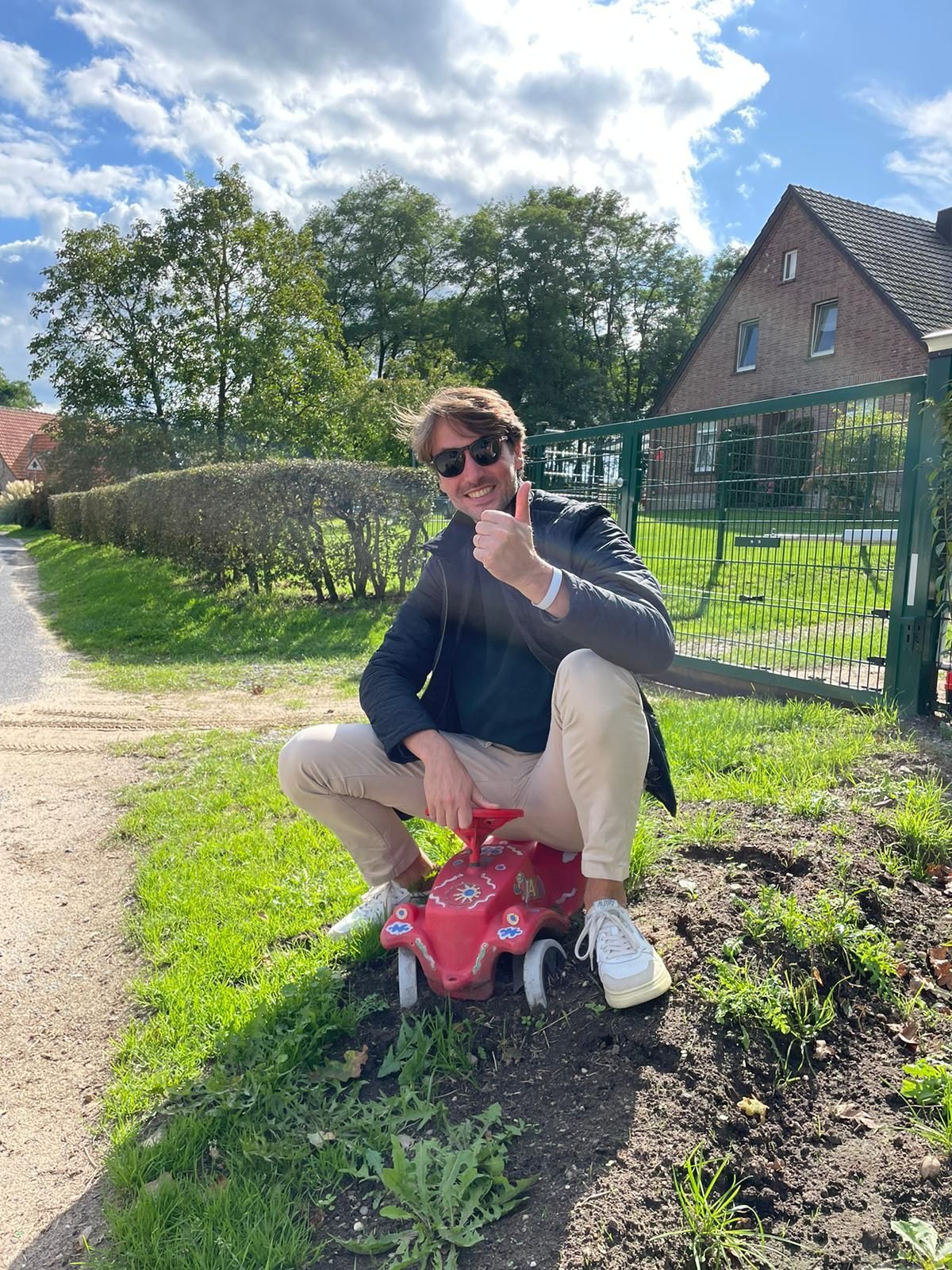 Een man steekt zijn duim omhoog terwijl hij gehurkt op een kleine rode auto in de buitenlucht zit.