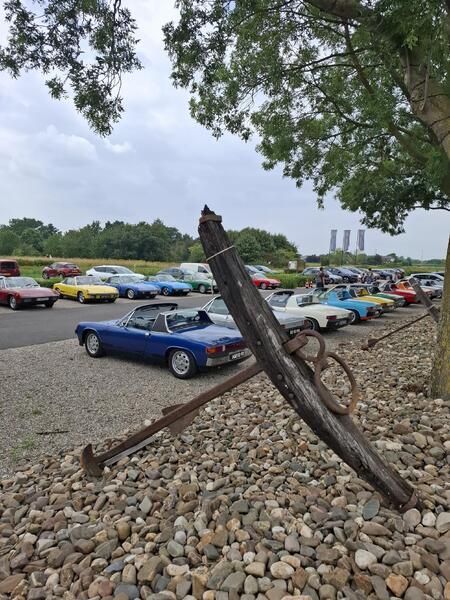 Een antiek anker voor een rij klassieke auto's geparkeerd op een grindpad, onder een bewolkte hemel.