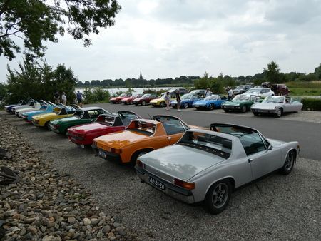Een rij klassieke Porsche 914's in verschillende kleuren staat geparkeerd langs een meer, onder een bewolkte hemel.
