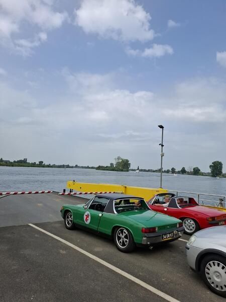 Groene en rode klassieke auto's op een veerboot, met water op de achtergrond, onder een bewolkte hemel.
