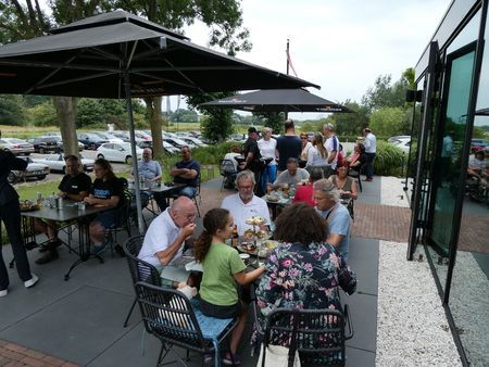 Een terrasje met mensen die aan tafels onder parasols zitten. Op de achtergrond staan ​​auto's geparkeerd.