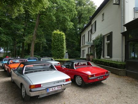 Er staan ​​diverse cabrioletversies van de Porsche 914 geparkeerd voor een wit gebouw met bomen.