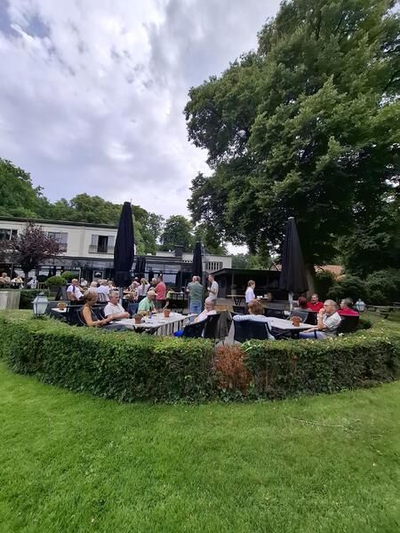 Mensen zitten aan tafels buiten op een terras, onder parasols, vlakbij een gebouw en groen.