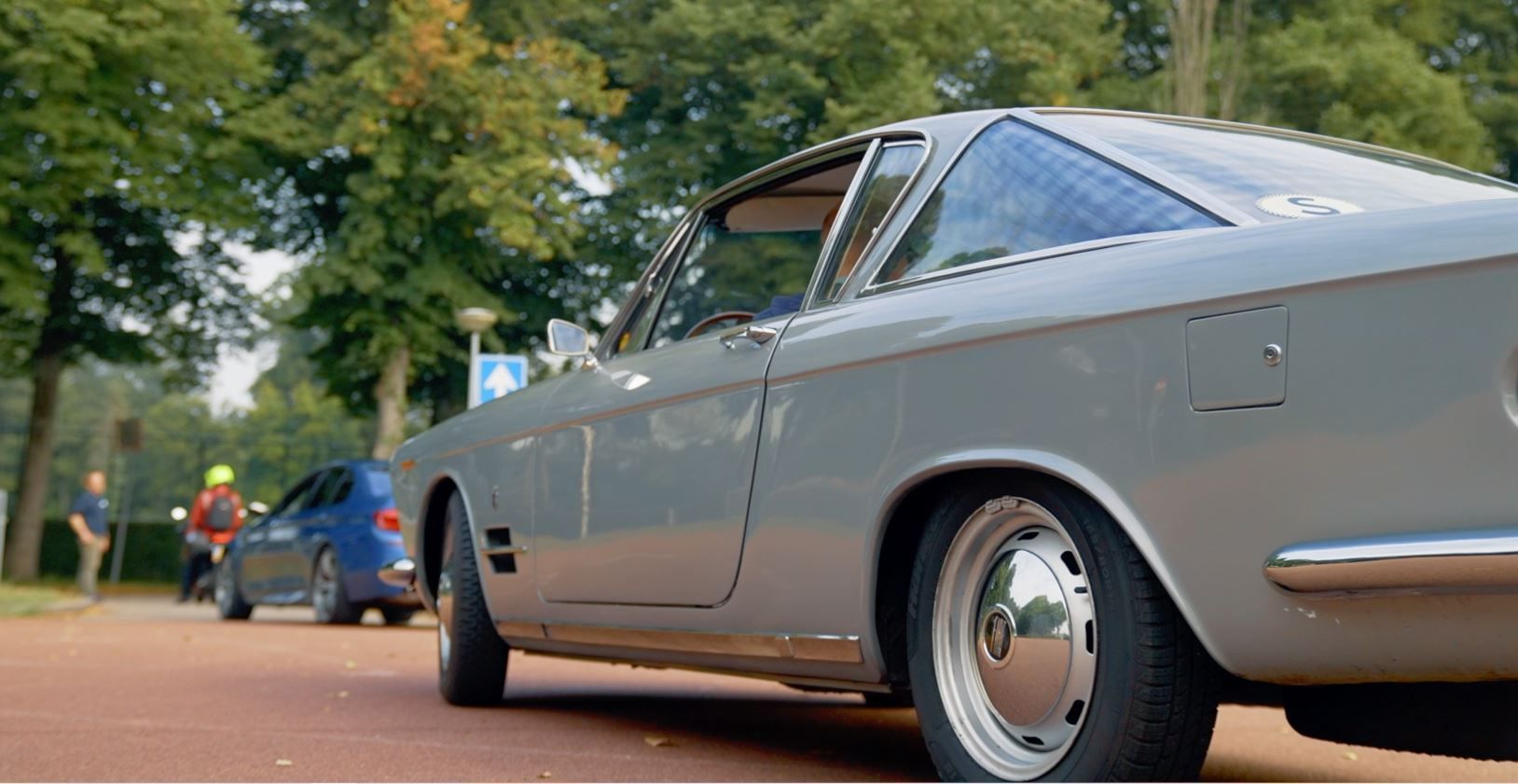 Een grijze oldtimer staat geparkeerd op een weg met bomen. Op de achtergrond zijn een andere blauwe auto en mensen te zien.