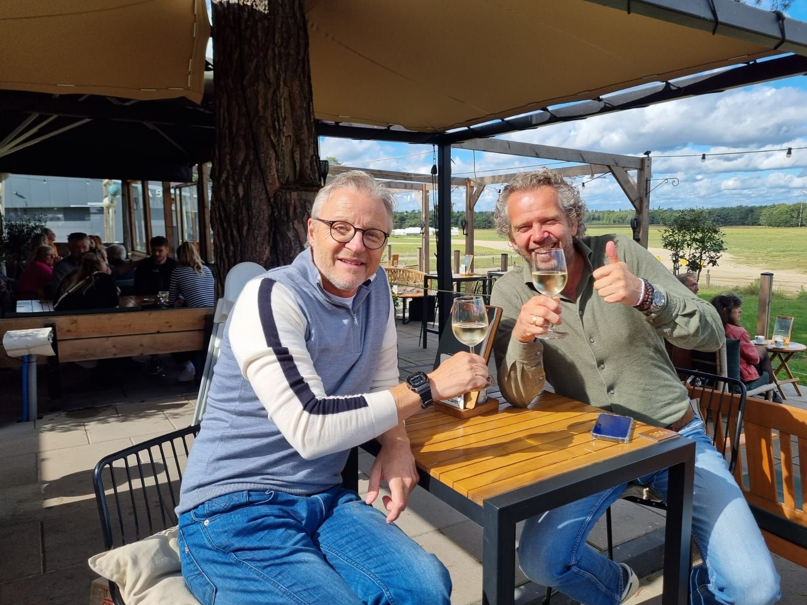 Twee mannen proosten buiten met wijn; zonnige dag, houten tafel. Een van de mannen glimlacht en steekt zijn duim omhoog.