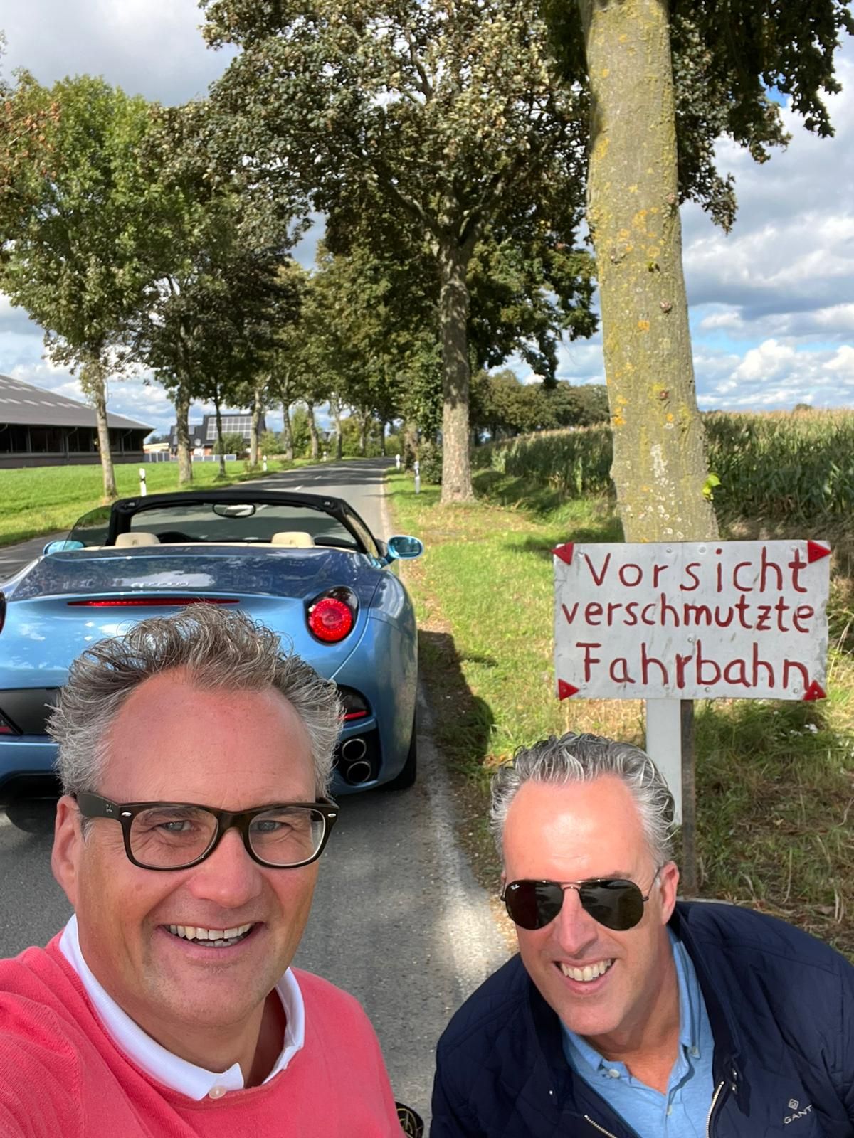 Twee mannen glimlachen voor een blauwe cabriolet; een bordje luidt: 