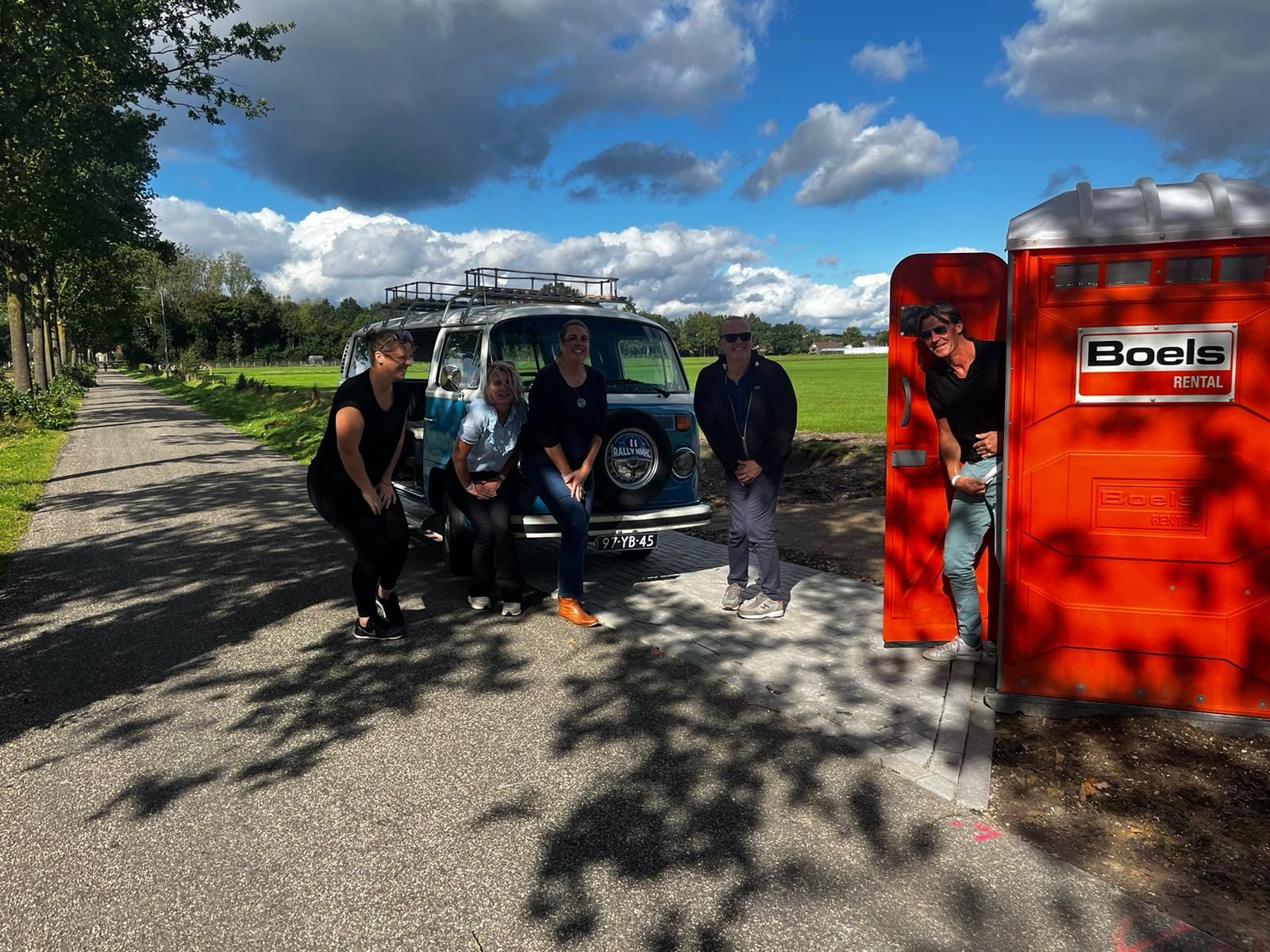 Een groep poseert bij een blauw busje vlakbij een oranje mobiel toilet op een zonnige dag.