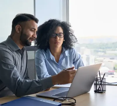 Two coworkers reviewing a laptop together at a bright office desk by a window.