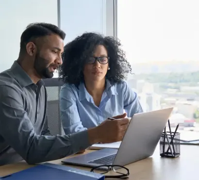 Two coworkers reviewing a laptop together at a bright office desk by a window.
