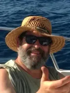 Smiling person in sunglasses and a straw hat giving a thumbs-up on a boat with blue water behind them