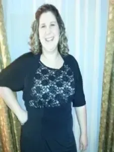 Woman in a black dress with lace bodice stands smiling against a light curtain backdrop.