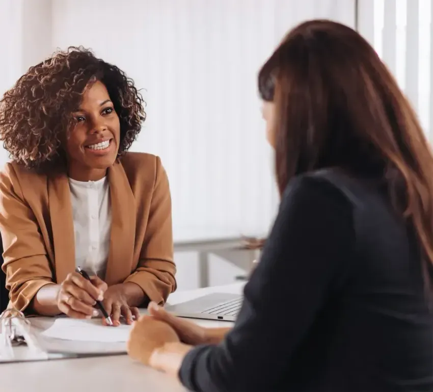 Two women in a bright office meeting, one smiling and taking notes across a desk.