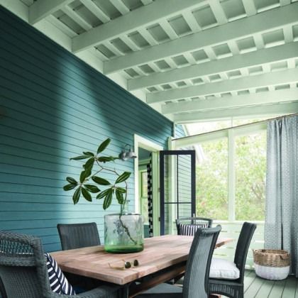 Modern patio dining area with teal shiplap wall, wooden table, and open glass doors to a bright garden