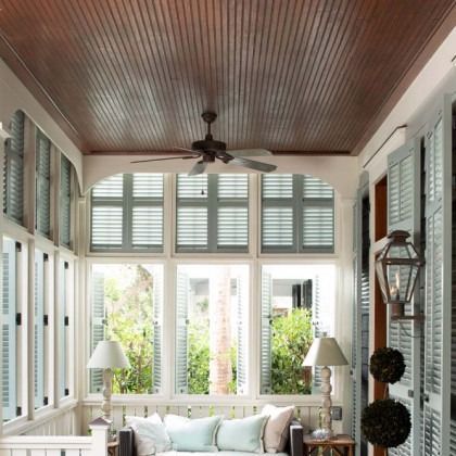 Bright sunroom with white shutters, a wood ceiling, ceiling fan, and light sofas by tall windows.
