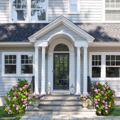 Front porch of a white house with columns, steps, potted flowers, and a black front door