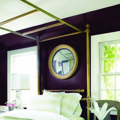 Purple bedroom with brass four-poster bed, white linens, round mirror, and bright windows.
