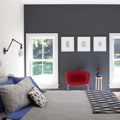 Modern bedroom with dark gray walls, red chair, patterned bed, and wall art between two windows
