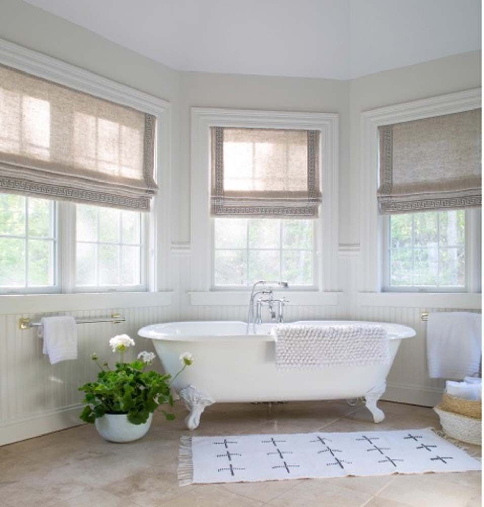 Bright white bathroom with a clawfoot tub, bay windows, woven shades, and a small potted plant.