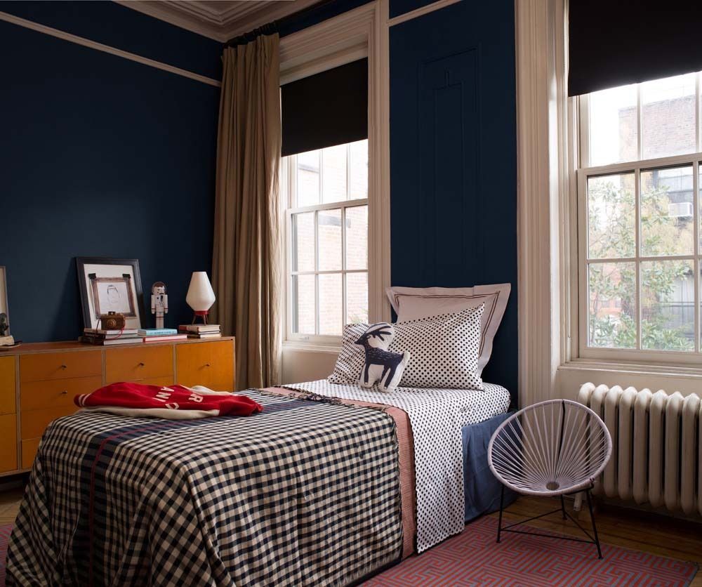Cozy bedroom with navy walls, plaid bedspread, wooden dresser, and large windows with beige curtains.
