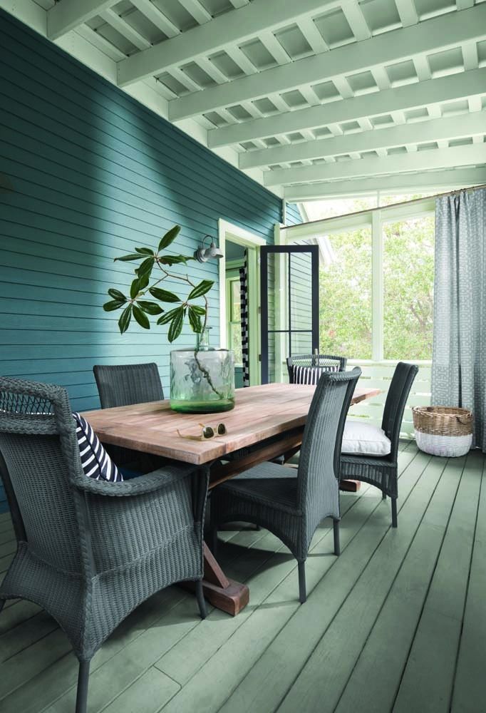 Covered patio with teal walls, wicker chairs, wooden table, and a vase of leafy branches by open doors.