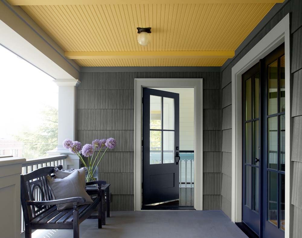 Covered porch with gray siding, yellow ceiling, black bench, flowers, and open doors to a bright interior