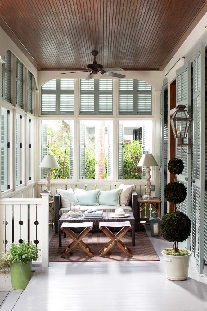 Bright sunroom porch with white shutters, wicker sofa, potted plants, and a ceiling fan