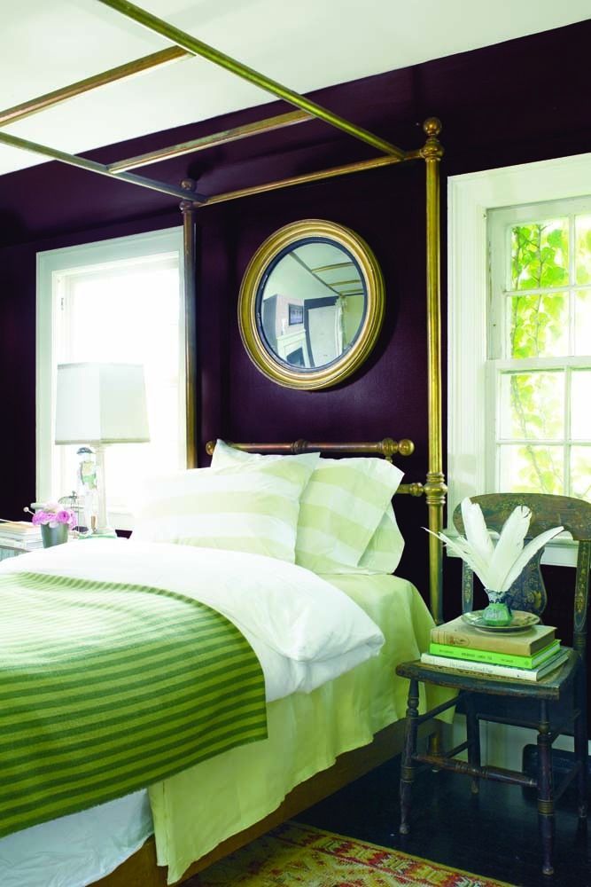 Purple bedroom with a four-poster bed, green bedding, and a round mirror between two windows
